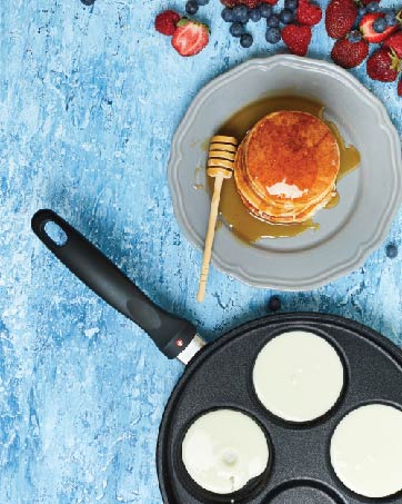 pancakes being made in a plett pan, with a plate of finished pancakes and berries nearby