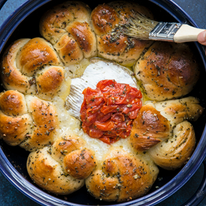 A photo of Garlic Knot Crown with Brie and Tomato Jam with someone brushing butter on the bread. 