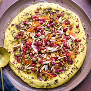 A photo of Jeweled Hummus with nuts and pomegranate seeds on a pink plate.