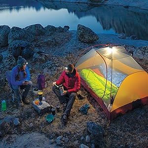 Two people camped out by some water sitting around their cooler at night. 